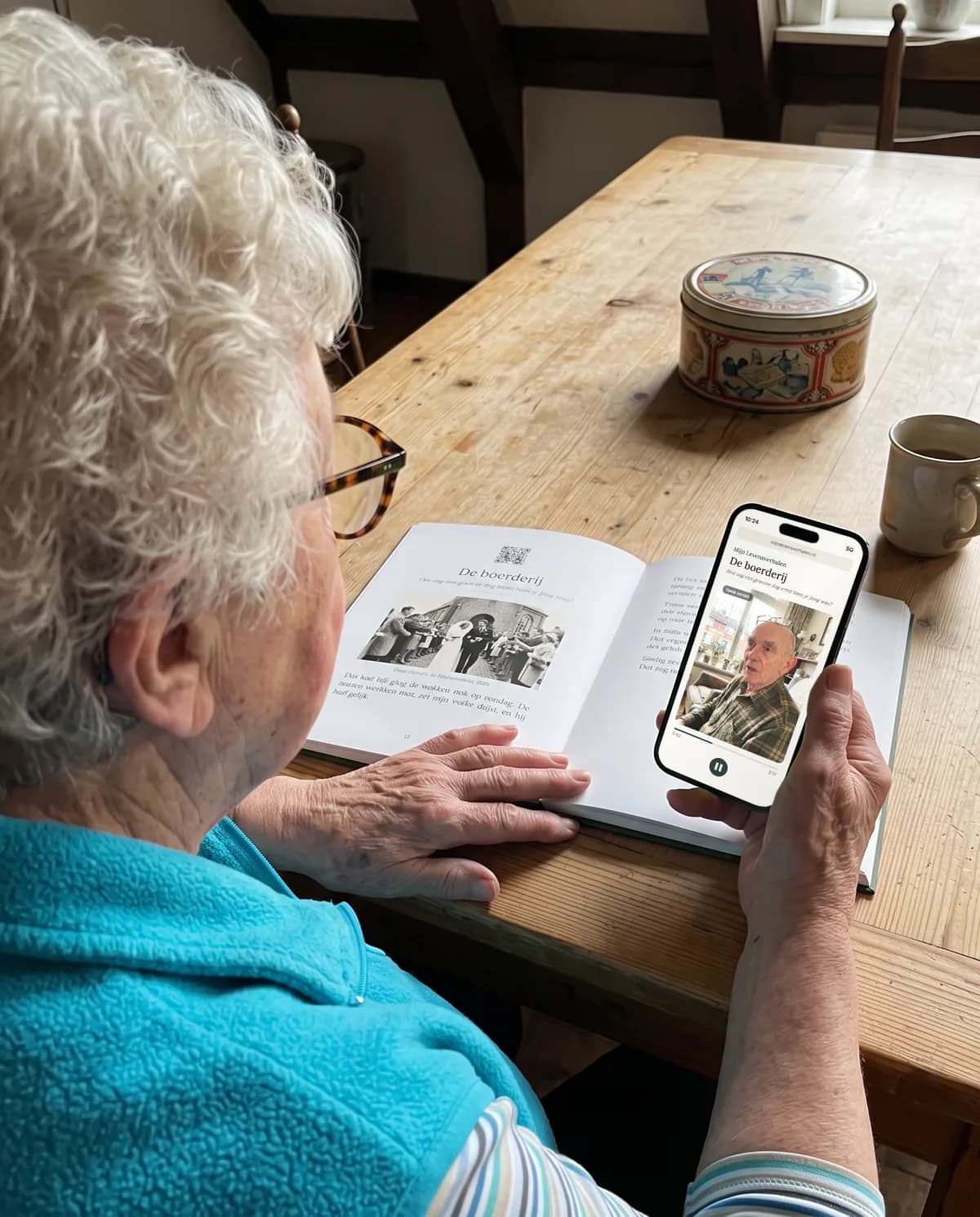 Een kleindochter luistert aan de keukentafel naar opa's verhaal op haar telefoon, met zijn boek ernaast.