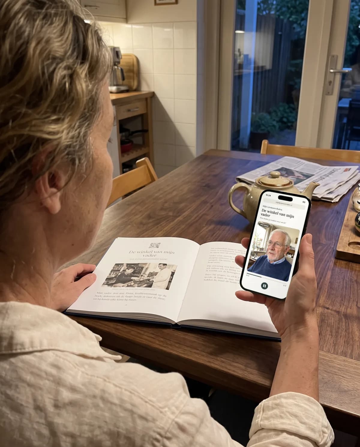 Een dochter met het boek van haar vader in de keuken, terwijl ze op haar telefoon naar zijn verhaal luistert.